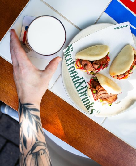Três baos de língua defumada num prato com a marca NiceFood&Drinks, ao lado de um chopp, sobre a mesa de azulejo da área externa.