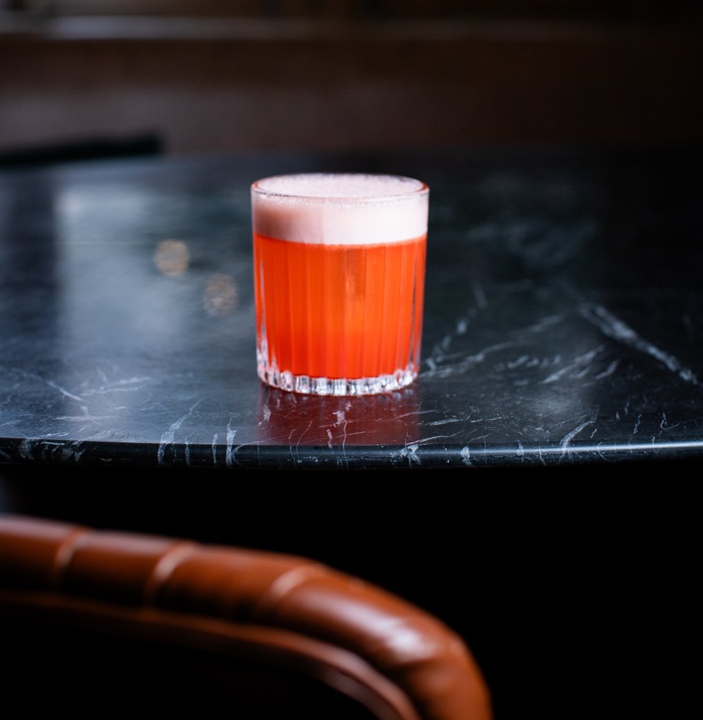 Um Aperol Sour numa taça canelada, sobre uma mesa de mármore preto, com o encosto de uma poltrona de couro caramelo em primeiro plano.