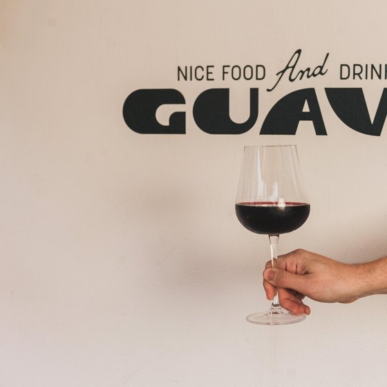Uma mão segurando uma taça de vinho tinto diante de uma parede clara onde está pintado “Nice Food And Drinks” sobre o nome Guava.