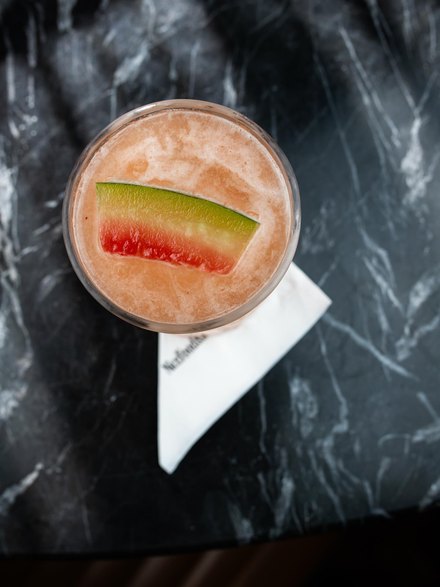 Drink Watermelon num copo alto, cor de melancia, com uma fatia de melancia na borda.